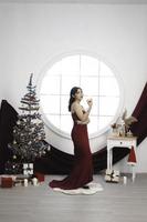 ritratto di un' bella giovane ragazza indossare un' rosso toga e potabile vino, sorridente a il telecamera, in piedi nel decorato Natale vivente camera in casa foto