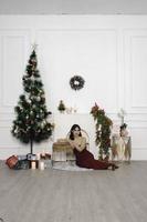 ritratto di bella giovane ragazza accogliente sedersi giù su il pavimento, sorridente indossare rosso toga nel decorato Natale vivente camera in casa foto