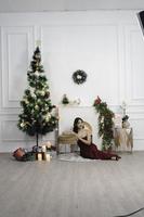 ritratto di bella giovane ragazza accogliente sedersi giù su il pavimento, sorridente indossare rosso toga nel decorato Natale vivente camera in casa foto