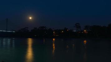 notte Visualizza con giallo Luna di il ponte al di sopra di il acquarico fiume Il prossimo per il città di Lago Agrio con luci di case illuminato tra il alberi foto