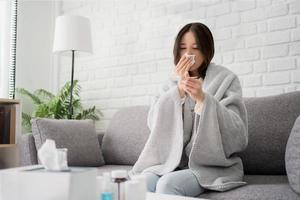 malato giovane asiatico donna sedersi sotto coperta su divano. femmina soffio naso, tosse, starnutire, dai un'occhiata Temp nel fazzoletto di carta a casa, sofferenza a partire dal influenza. foto