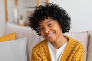 bellissimo africano americano ragazza con afro acconciatura sorridente seduta su divano a casa interno. giovane africano donna con Riccio capelli ridendo. la libertà felicità spensierato contento persone concetto. foto