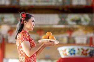 asiatico donna nel rosso cheongsam qipao vestito è offerta mandarino per il ancestrale Dio dentro Cinese buddista tempio durante lunare nuovo anno per migliore desiderio benedizione e bene fortuna concetto foto