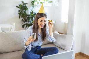 virtuale festa. contento giovane donna nel cappello avendo video conferenza in linea incontro con amici e famiglia, Tenere bicchiere di vino, tostatura e festeggiare compleanno, soggiorno a casa foto