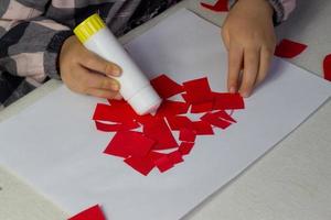 poco ragazza fabbricazione rosso cuore applique con carta e colla su bianca foglio, san valentino giorno saluto carta, vacanza regalo per madri giorno, mano fatto bambini presente, amore simbolo con carta, bambino attività foto