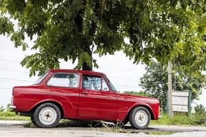 un' Visualizza di il vecchio classico rosso berlina parcheggiata nel il pioggia per un' lungo tempo sotto il frondoso verde alberi. foto