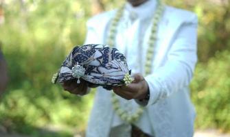 bellissimo sposo Tenere tradizionale copricapo mentre indossare nozze vestito con gelsomino e magnolia fiore collana per un' tradizionale nozze cerimonia nel Indonesia foto