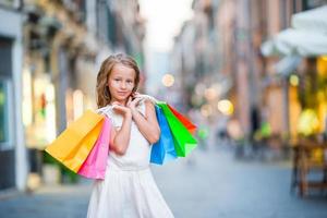 ritratto di adorabile poco ragazza a piedi con shopping borse all'aperto nel Roma. moda bambino piccolo ragazzo nel italiano città con sua shopping foto