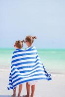 adorabile poco ragazze avvolto nel asciugamano a tropicale spiaggia dopo nuoto nel il mare foto