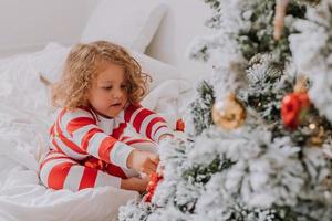 poco ragazza nel Natale a strisce pigiama decora il Natale albero. stile di vita. alto qualità foto