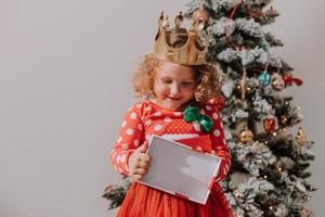 poco Riccio ragazza nel un' rosso carnevale vestito con un' Santa Stampa e un' corona è giocando con un' giocattolo cervo Il prossimo per il Natale albero. stile di vita, figli di emozioni. spazio per testo. alto qualità foto