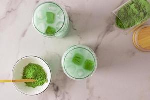 Due bicchieri di matcha latte macchiato ghiaccio tè e ghiaccio cubi su un' bianca marmo sfondo. un' ciotola di verde tè polvere e un' bambù misurazione cucchiaio. superiore Visualizza. foto