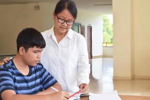 giovane asiatico ragazzo è fare il suo Incarico e ascoltando per il suo femmina anziano insegnanti consigli di scuola progetto, adulto aiuta bambini per fare scuola progetto concetto. foto
