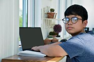 giovane asiatico ragazzo si siede nel davanti di finestra di vetro e utilizzando il suo il computer portatile per opera in linea e ascoltando per musica, ricreativo attività e raccolta adolescenti concetto. foto