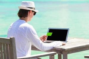 giovane uomo Lavorando su il computer portatile con credito carta durante tropicale vacanza foto