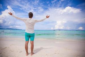 indietro Visualizza di giovane uomo diffusione il suo braccia in piedi su bianca sabbioso spiaggia foto