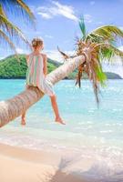 adorabile poco ragazza seduta su palma albero durante estate vacanza su bianca spiaggia foto