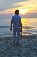 giovane uomo con un' bottiglia di vino e bicchieri su il spiaggia a tramonto foto