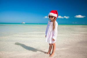 poco ragazza nel rosso cappello Santa Claus e occhiali da sole su il spiaggia foto