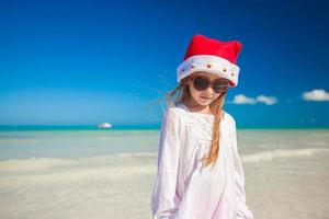 poco ragazza nel rosso cappello Santa Claus e occhiali da sole su il spiaggia foto