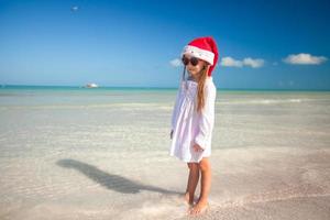 poco ragazza nel rosso cappello Santa Claus e occhiali da sole su il spiaggia foto