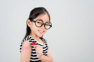 asiatico poco ragazza indossare bicchieri mostrando sua braccio con bendare dopo virus vaccino contento ragazzo e bambino vaccinazione campagna. proteggere il tuo ragazzo con vaccino concetto foto