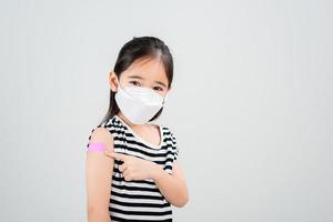 asiatico poco ragazza con medico maschera mostrando sua braccio con bendare dopo virus vaccino. ragazzo e bambino vaccinazione campagna. proteggere il tuo ragazzo con vaccino concetto foto