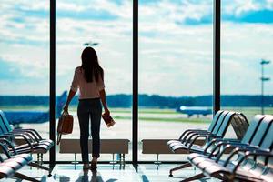 giovane donna nel internazionale aeroporto con sua bagaglio sfondo grande finestra. linea aerea passeggeri nel un aeroporto sala in attesa per volo aereo foto