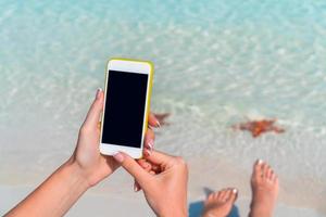 avvicinamento cellulare nel femmina mani su il sfondo di turchese oceano a tropicale spiaggia foto