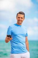 giovane uomo Tenere mobile Telefono nel il suo mano mentre a piedi su il spiaggia foto