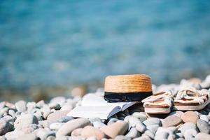 spiaggia cappello su ha aperto libro con protezione solare e scarpe su ciottolo spiaggia foto