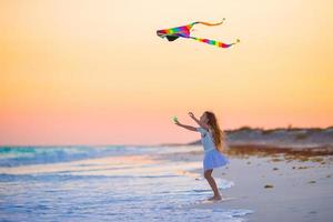 poco ragazza con volante aquilone su tropicale spiaggia a tramonto. ragazzo giocare su oceano costa. bambino con spiaggia giocattoli. foto