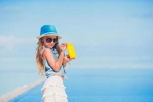 poco ragazza con bottiglia di sole crema seduta su il bordo di nuoto piscina foto