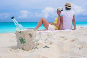 bottiglia di bianca vino e Due bicchieri sfondo contento coppia su sabbioso spiaggia foto