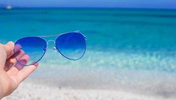 vicino su di colorato blu occhiali da sole nel mano su tropicale spiaggia foto