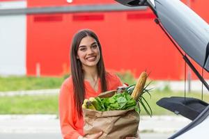 caucasico brunetta andando Tenere carta borse con cibo prodotti. giovane donna mettendo pacchetto con drogheria e verdure in auto tronco. attraente caucasico femmina shopping nel centro commerciale o drogheria memorizzare foto