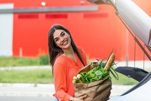 caucasico brunetta andando Tenere carta borse con cibo prodotti. giovane donna mettendo pacchetto con drogheria e verdure in auto tronco. attraente caucasico femmina shopping nel centro commerciale o drogheria memorizzare foto