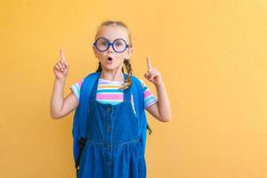 emotivo eccitato sorpresa viso scolara bambino nel bicchieri nel scuola blu uniforme con zaino scuola Borsa Aperto bocca punto dita su per vuoto copia spazio attento isolato su giallo sfondo foto