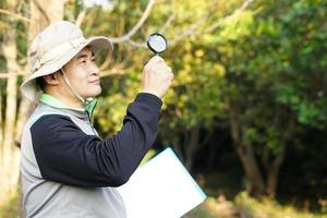 bello asiatico uomo botanico è a foresta per sondaggio botanico impianti, detiene carta appunti e ingrandimento bicchiere. concetto, esaminare, Esplorare, ricerca, studia di ambiente, impianti e natura. foto