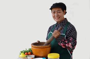 contento asiatico anziano donna è cucinando, indossare capocuoco berretto e grembiule, detiene pestello, mortaio e piatto di peperoncini. concetto, cucinando per famiglia. tailandese cucina stile di vita. anziano attività. foto