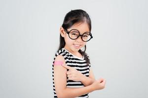 asiatico poco ragazza indossare bicchieri mostrando sua braccio con bendare dopo virus vaccino contento ragazzo e bambino vaccinazione campagna. proteggere il tuo ragazzo con vaccino concetto foto