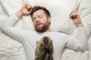 uomo nel bianca pigiama è addormentato, e il suo adorabile gatto bugie su il suo il petto. superiore Visualizza. foto