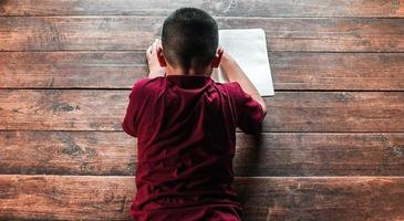 ragazzo seduta su il di legno pavimento scrittura un' libro o fare compiti a casa a casa. superiore Visualizza di ragazzo scrittura libro foto