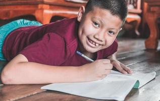 avvicinamento di un asiatico ragazzo seduta su il di legno pavimento scrittura un' libro o fare compiti a casa con un' sorridente divertimento viso a casa. foto