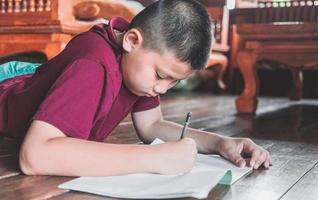 avvicinamento di un asiatico ragazzo seduta su il di legno pavimento scrittura un' libro o fare compiti a casa con un' molto determinato viso a casa. foto