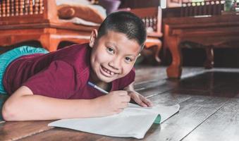 avvicinamento di un asiatico ragazzo seduta su il di legno pavimento scrittura un' libro o fare compiti a casa con un' sorridente divertimento viso a casa. foto