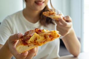 un asiatico donna è mangiare un' Pizza lui ordinato. durante il rompere, godere italiano pezzi con delizioso formaggio a casa. foto