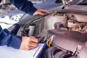 mani di giovane professionale meccanico nel uniforme scrittura su appunti contro auto nel Aperto cappuccio a il riparazione box auto. Manutenzione se. auto servizio e Manutenzione foto