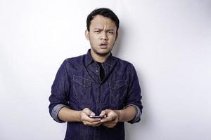 un' riflessivo giovane asiatico uomo è indossare blu camicia Tenere il suo Telefono e sembra confuso, isolato di bianca sfondo foto