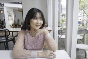 un' sorridente asiatico donna indossare rosa vestito mentre seduta vicino finestra foto